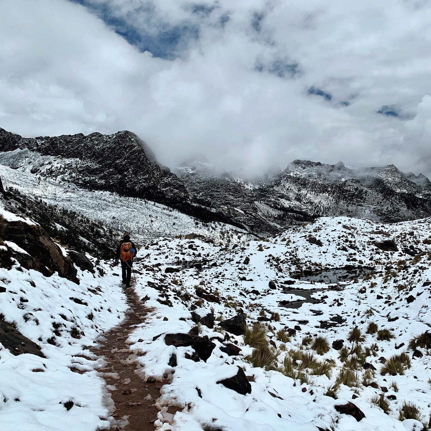 A distant figure walks away from the camera along a narrow dirt path through snow-covered mountains.